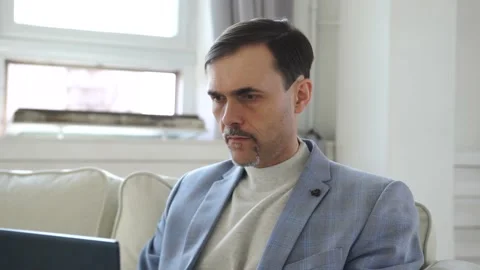 A man with a beard sits in a jacket on the sofa, typing on a laptop keyboard Stock Footage 152473565