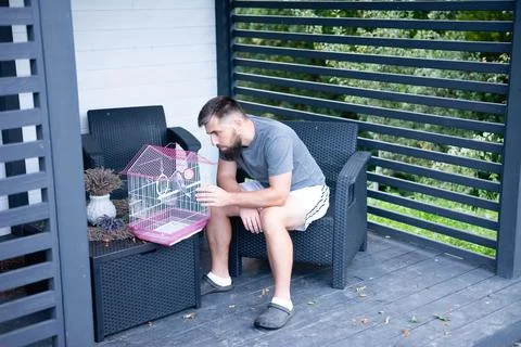 Man with beard sits outside, looking at a bird in a cage. Foto stock