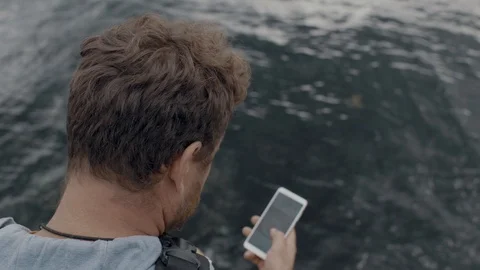 Man with a beard sits on a pier in the sea and looks at the phone, clicking Stock Footage 115480483