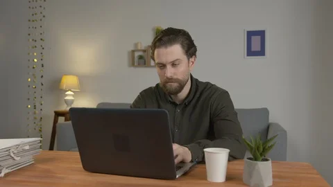 A man with a beard sits at a table, works in a laptop, at home, drinks hot 스톡 동영상 129082189