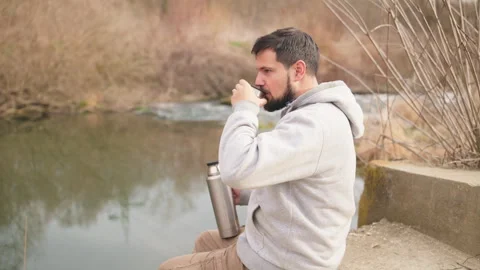 A man with beard is sitting on the edge of a river drinking from a thermos Stock Footage 265761143