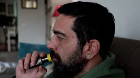 Man with beard sitting on the sofa at home smokes an electronic cigarette. Stock Footage 233914609