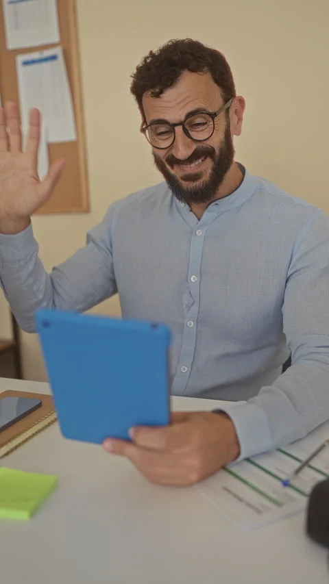 Man with beard smiling and waving at tablet during video call in office set.. Stock Footage 311938672