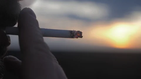 Man with a beard smoking a cigarette at sunset. Hand with burning cigarette