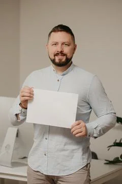 Man with a beard is standing with an empty sheet of white paper near a comput Stock Photos