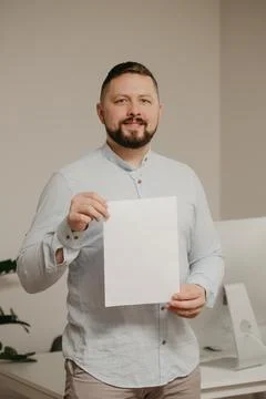 Man with a beard is standing with an empty sheet of white paper near a comput Stock Photos