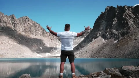 Man with a beard stands on the top of a mountain by a lake and spreads his arms Stock Footage 138445913