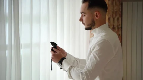 A man with a beard stands by the window in a white shirt and examines a bow tie Video stock 131339974