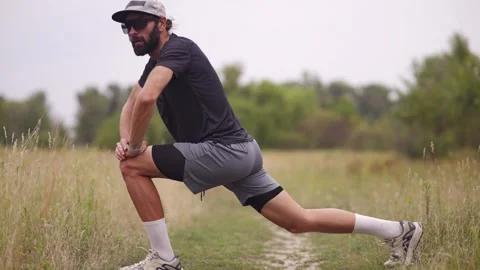 Man with beard stretching in forward lunge on grassy trail wearing hat outdoors Stock Footage 313951532