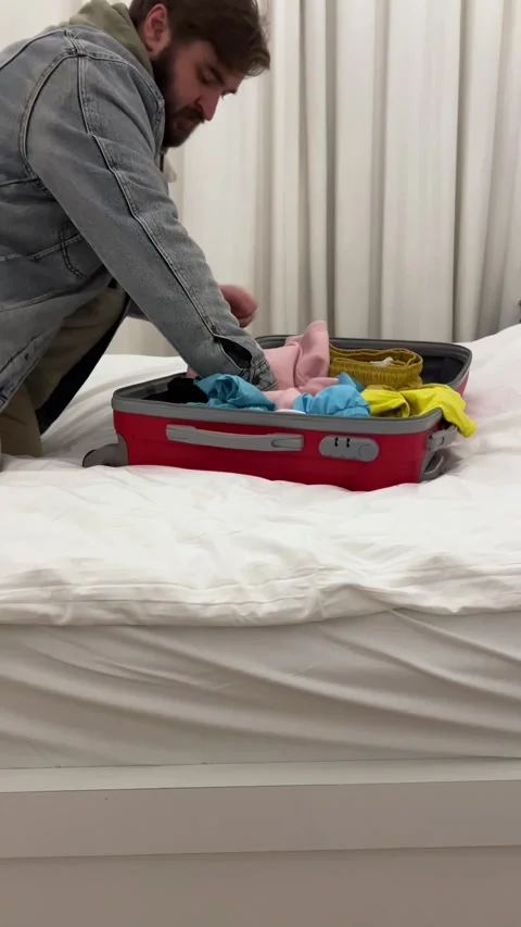 Man with beard throws clothes into red suitcase in hotel bed room Stock Footage 306262017