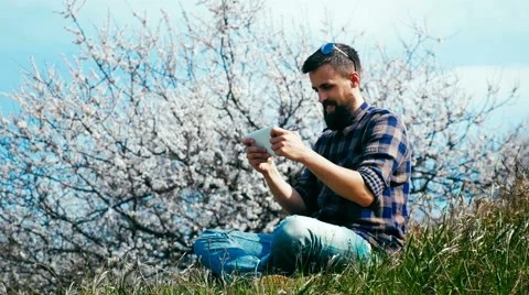 Man with a beard uses a tablet on nature near blossoming tree Stock Footage 62439386