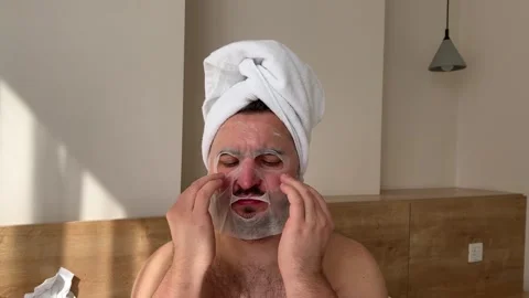 Man with beard, white towel on head, bare shoulders is put on cosmetic face mask Stock Footage 302767803