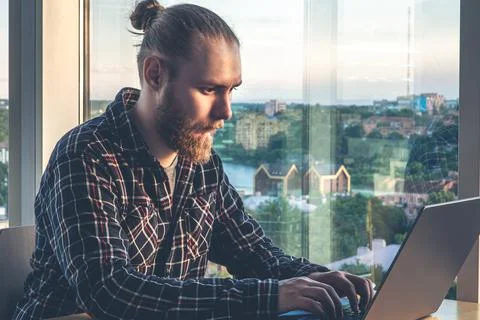 A man with a beard works at a computer, sitting in the office by the window. Stock Photos