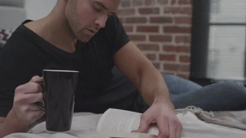 Man on bed reading a book and drinking coffee Stock Footage 70808139