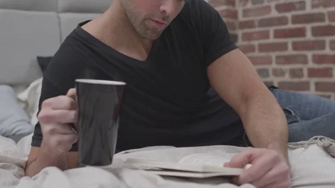 Man on bed reading a book and drinking coffee Stock Footage 70808393