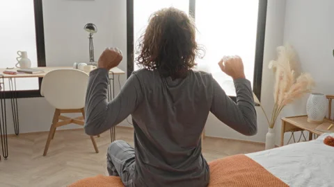Man on bed sits at edge and stretches both arms upward toward window with d.. Stock Footage 337377133