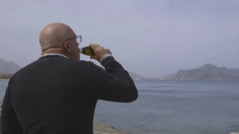 Man from behind drinks beer on a beach while looking at the sea. Video stock 305243369