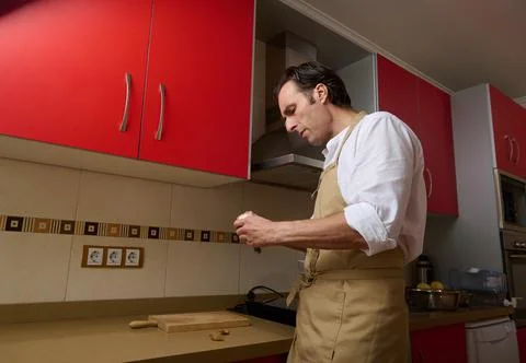 Man In Beige Apron Preparing Dinner In A Modern Kitchen With Red Cabinets Foto stock