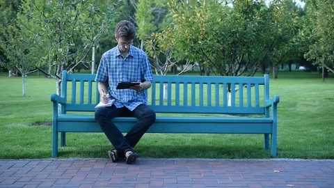 Man on bench in park using a tablet and writing in notepad Stock Footage 71313593