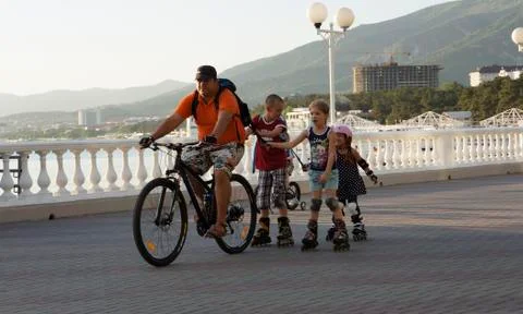 A man on a bicycle riding with children on rollers along the waterfront Stock Photos