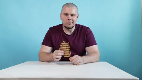 A man biting into a grilled sandwich while sitting at a table, wearing a ma.. Stock Footage 298236820
