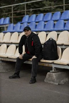 Man in black bomber jacket and cap sits alone in empty stadium tribune with.. Фото