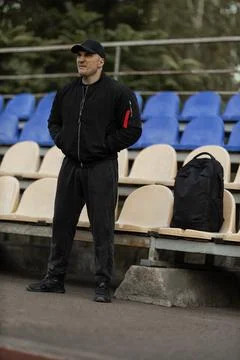 Man in black bomber jacket and cap watches intense sports game from empty stands Stock Photos
