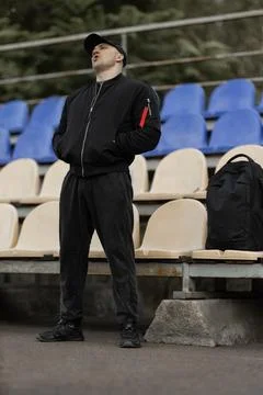 Man in black bomber jacket and cap reacts among empty stands during a sports Stock Photos