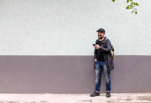 A man in a black cap, light summer raincoat and glasses holds a phone in his Stock Photos