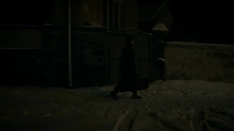 A man in a black cloak and hat walks along a wall on the sidewalk. 動画素材 102139417