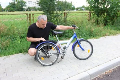 Man in black clothes checking broken chain on a kids bicycle on sidewalk near Foto stock