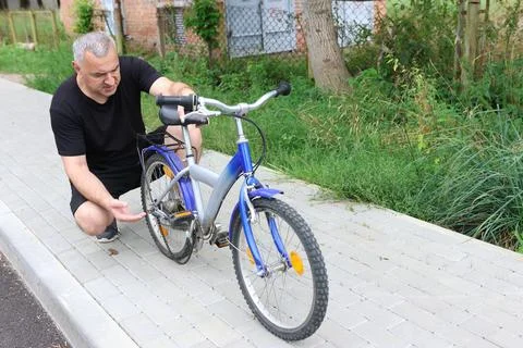 Man in black clothes checking broken chain on a kids bicycle on sidewalk near Stock Photos