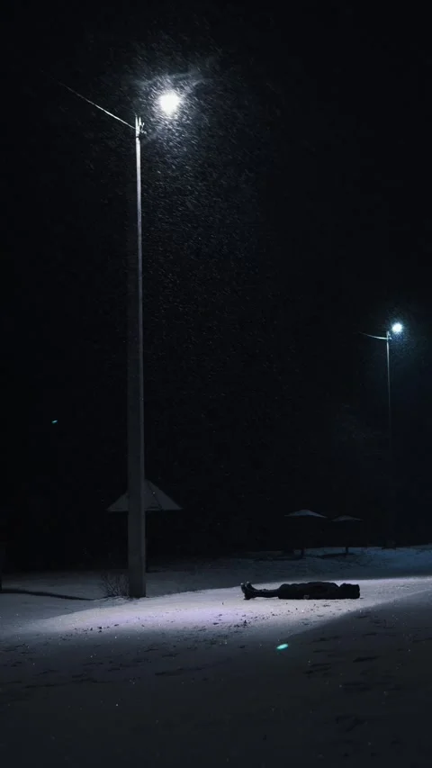 A man in a black down jacket lies on his back at night in the snow under a lante Stock Footage 326324913