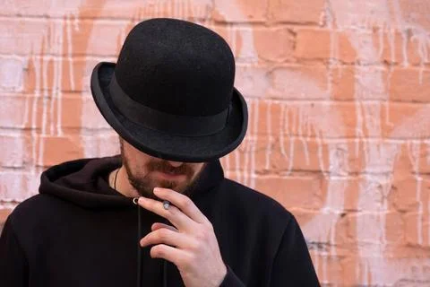A man in a black hat smoking a cigarette outdoors against a brick wall Stock Photos