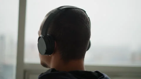 Man in black headphones facing blurred cityscape window indoors featuring Stock Footage 346652324