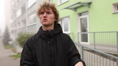 A man in a black hooded jacket stands near a residential building, looking upwar Stock Footage 290185554