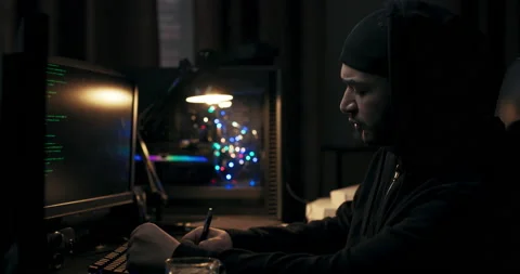 Man in black jacket sits at computer desk in dark room. Guy with beard holds pen Stock Footage 214699876