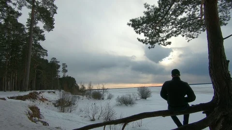 A man in a black jacket, sitting on the bank of a frozen river in the evening Video stock 87462600