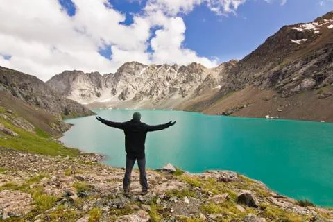 Man in black jacket standing with hands-up above blue lake in mountains Stock Photos