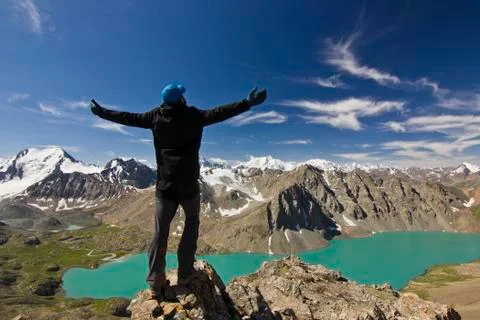 Man in black jacket standing with hands-up above blue lake in mountains Stock Photos