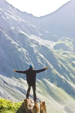 Man in black jacket standing on stone with hands-up above mountais at sunset Stock Photos