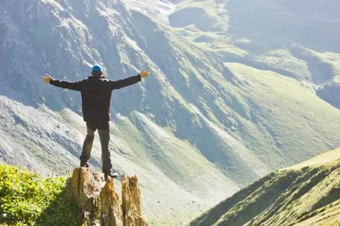 Man in black jacket standing on stone with hands-up above mountais at sunset Stock Photos