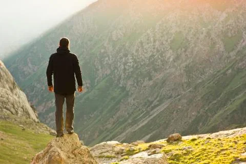 Man in black jacket standing on stone with hands-up above mountais at sunset Stock Photos