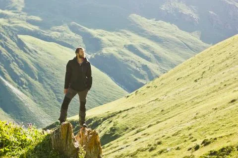 Man in black jacket standing on stone with hands-up above mountais at sunset Stock Photos