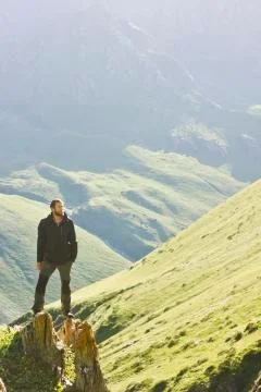 Man in black jacket standing on stone with hands-up above mountais at sunset Stock Photos