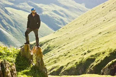 Man in black jacket standing on stone with hands-up above mountais at sunset Stock Photos