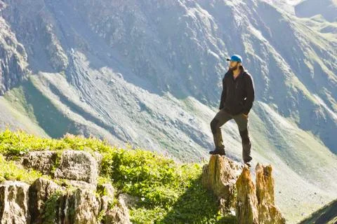Man in black jacket standing on stone with hands-up above mountais at sunset Stock Photos