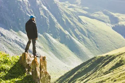 Man in black jacket standing on stone with hands-up above mountais at sunset Stock Photos