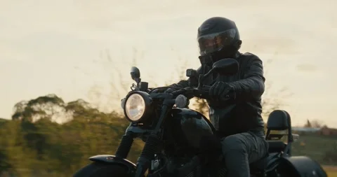 Man in a black leather jacket driving a black motorcycle through the dusk sky. Video stock 242175957
