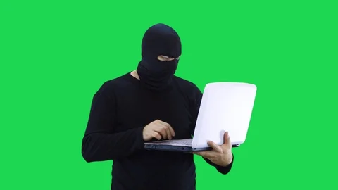 A man in a black mask enters data into a laptop, on a green background Stock Footage 127934643